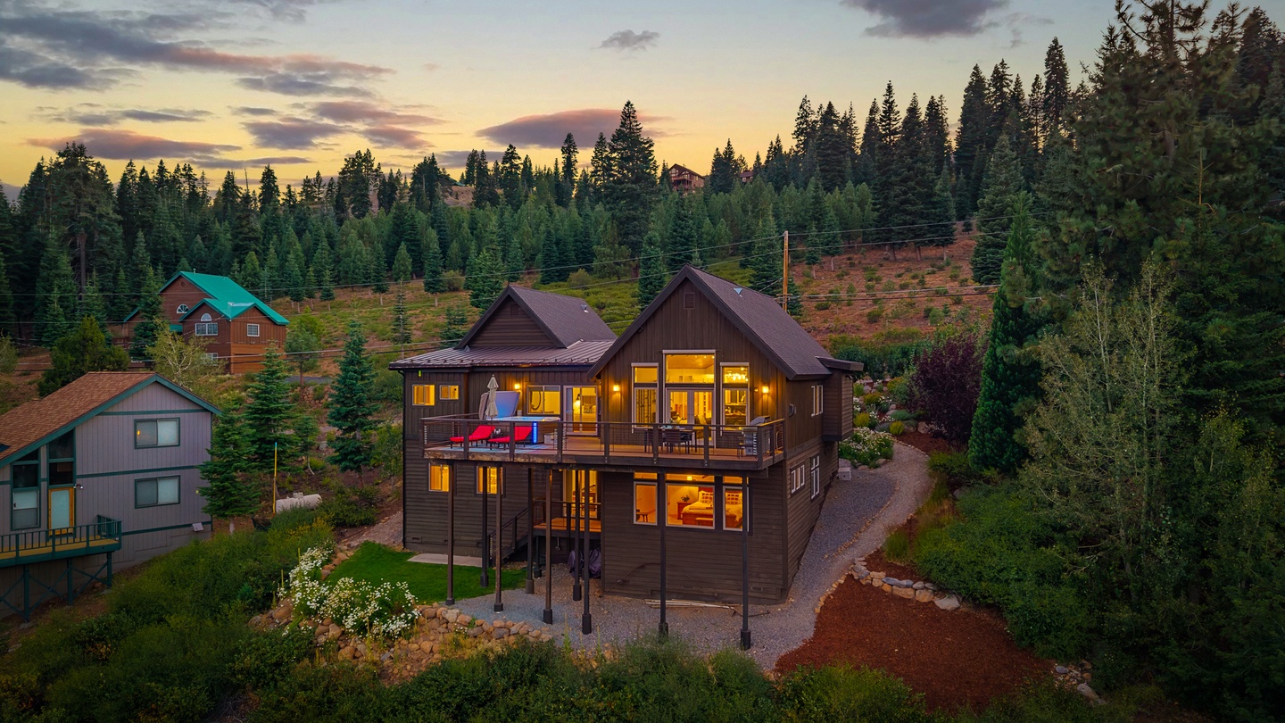 An aerial view captures this mountain retreat at twilight, nestled among towering evergreens with a dramatic sunset painting the sky. The property showcases multi-level outdoor decks and warm interior lighting in a forested hillside setting.