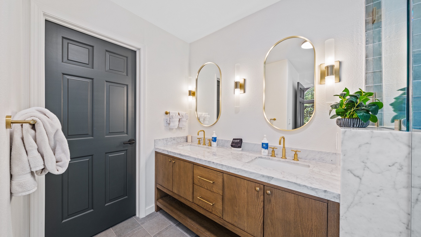 Elegant bathroom with dual marble vanities, gold fixtures, and oval mirrors. Soft sconces and a luxurious glass-walled shower create the perfect retreat.