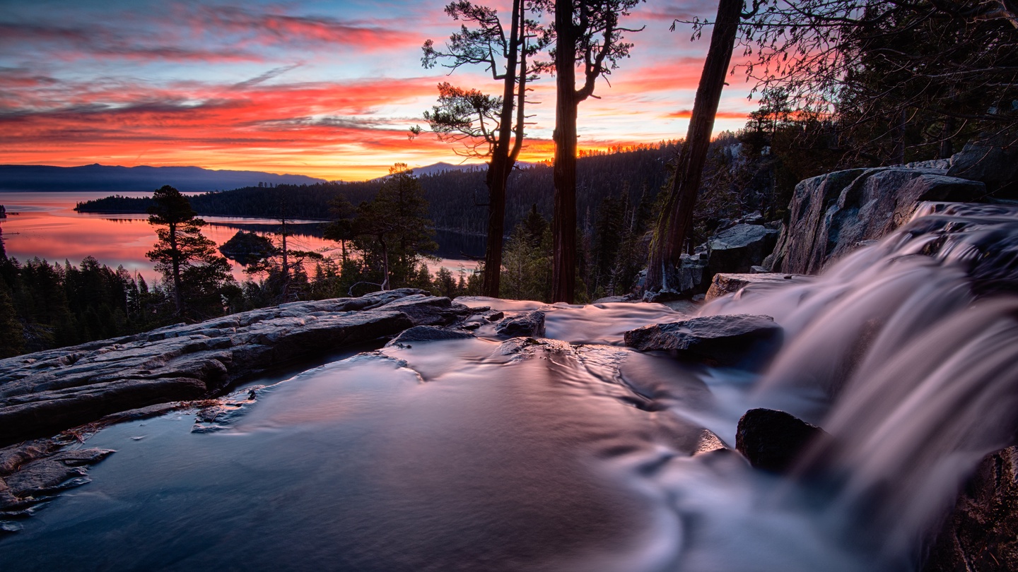 Stunning waterfall cascades through rocky terrain as vibrant sunset paints the sky over distant lake waters and forested mountains.
