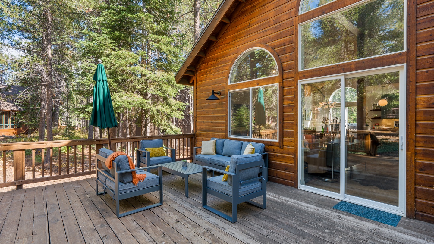 Charming rustic cabin exterior nestled among tall pines, featuring a spacious deck where you can unwind with comfortable seating, soak in the forest surroundings, and seamlessly step inside through the bright glass doors.