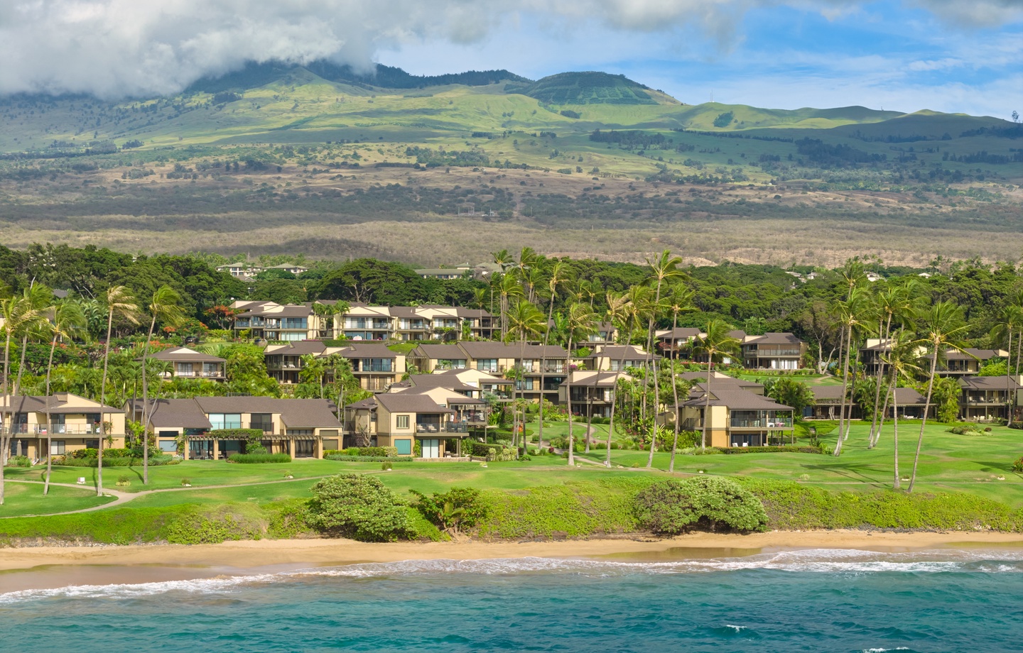 Aerial view of a beachfront resort community nestled between turquoise waters and dramatic mountain landscapes, surrounded by palm trees and lush golf course grounds.