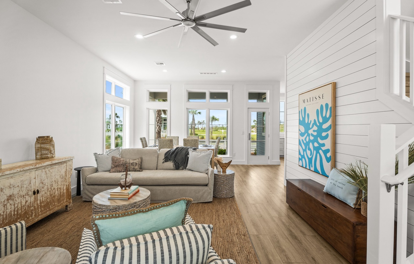 Sun-filled retreat with crisp white shiplap walls, soft coastal touches, and plush seating. Large windows frame views of the palm-dotted landscape beyond.