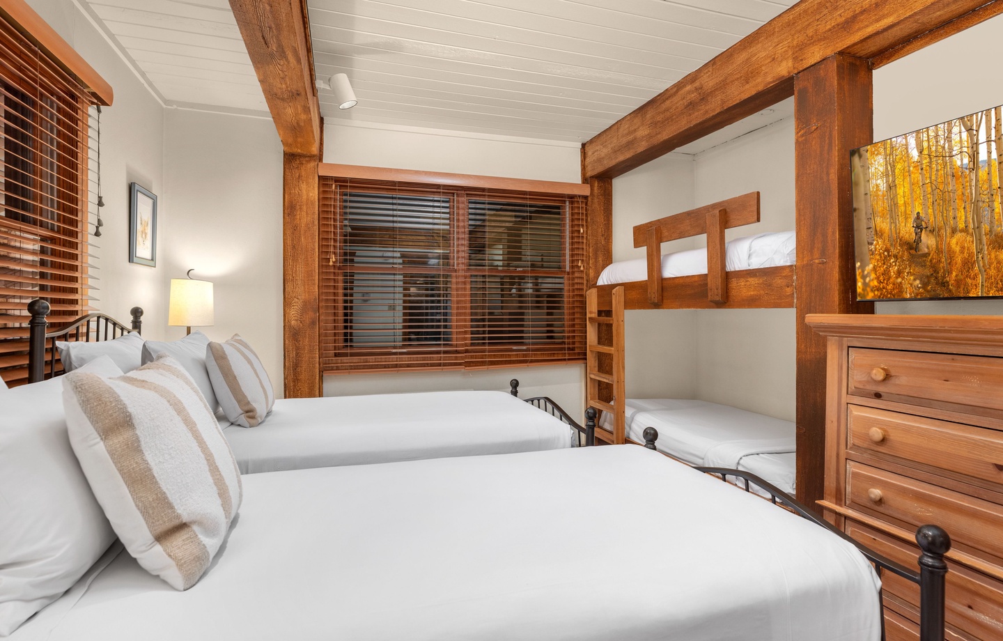 Your cozy bedroom blends rustic wooden beams and warm white linens, with sleeping options for families and soft natural light filtering through wooden blinds.