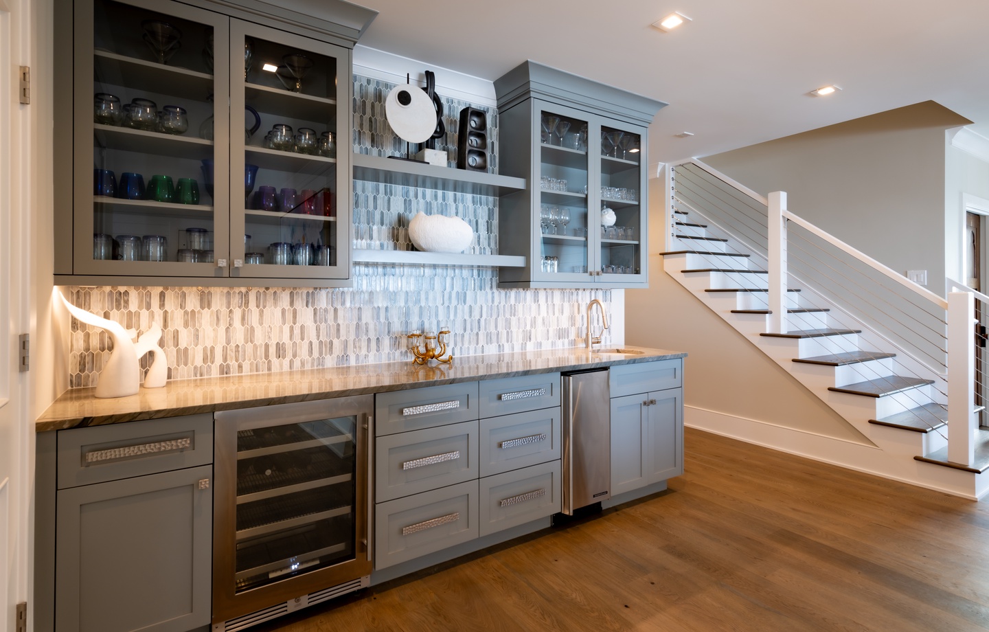 - Elegant gray cabinetry with glass front doors adds a refined touch to this wet bar - Features a wine fridge, sink, and plenty of drawers for effortless entertaining - Textured backsplash and warm wood floors blend style and function
