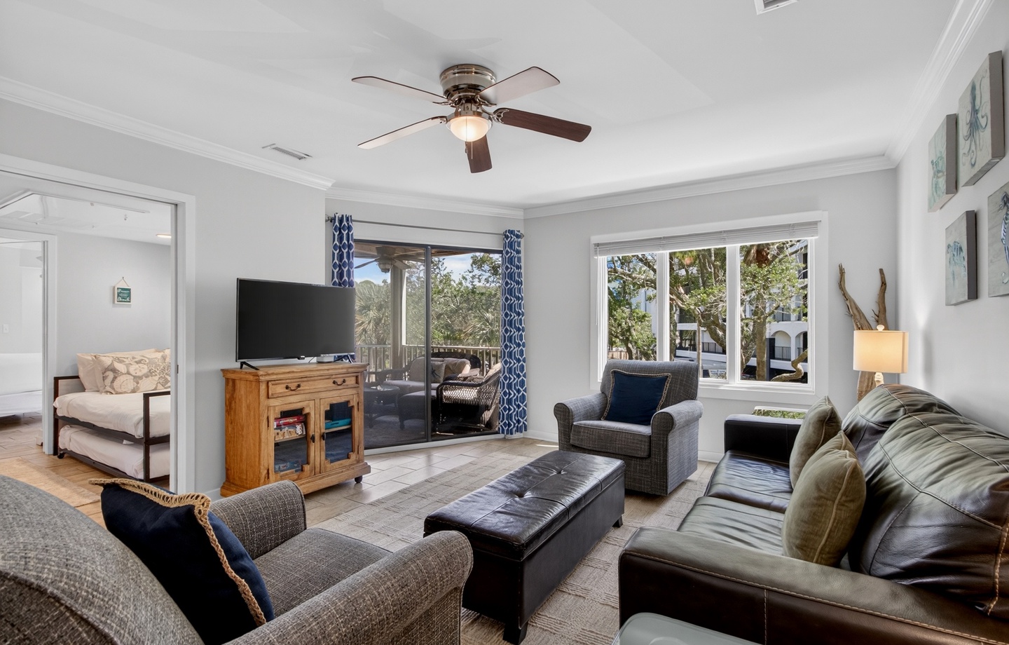 Unwind in this comfortable living room, where you can enjoy coastal breezes through the ceiling fan and abundant natural light from expansive windows framing lush greenery outside.