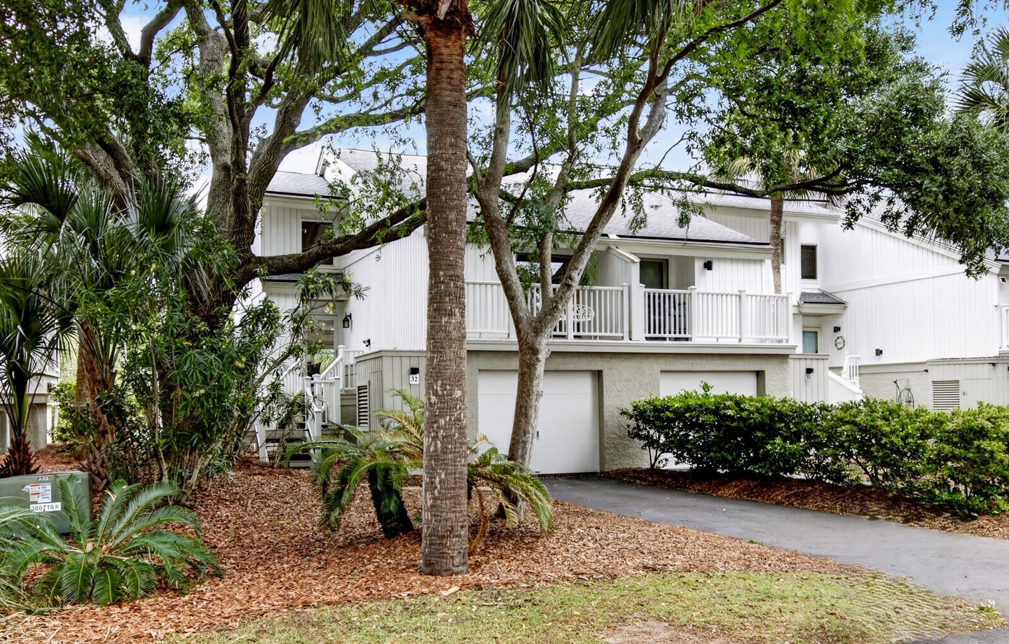 A coastal retreat nestled among lush tropical landscaping with palms and live oaks, featuring a private driveway leading to garage parking beneath covered balconies.