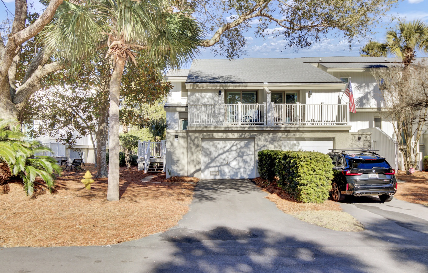 Charming two-story beach house with tropical palms and spacious balcony, featuring convenient parking and welcoming American flag display.
