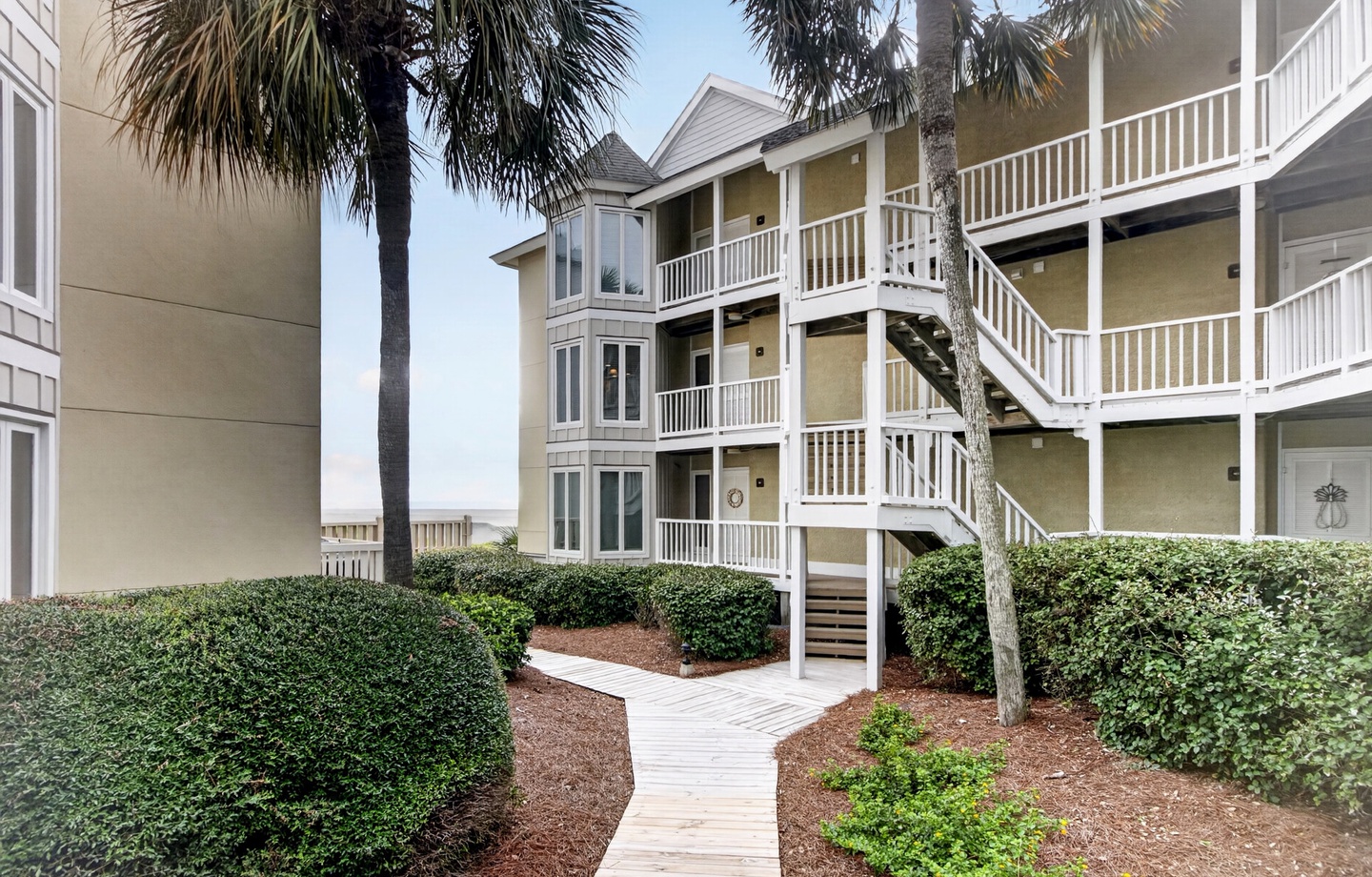 Coastal property buildings with white-railed balconies framing a sunny courtyard walkway, lined by palm trees and manicured hedges—your vacation getaway awaits.