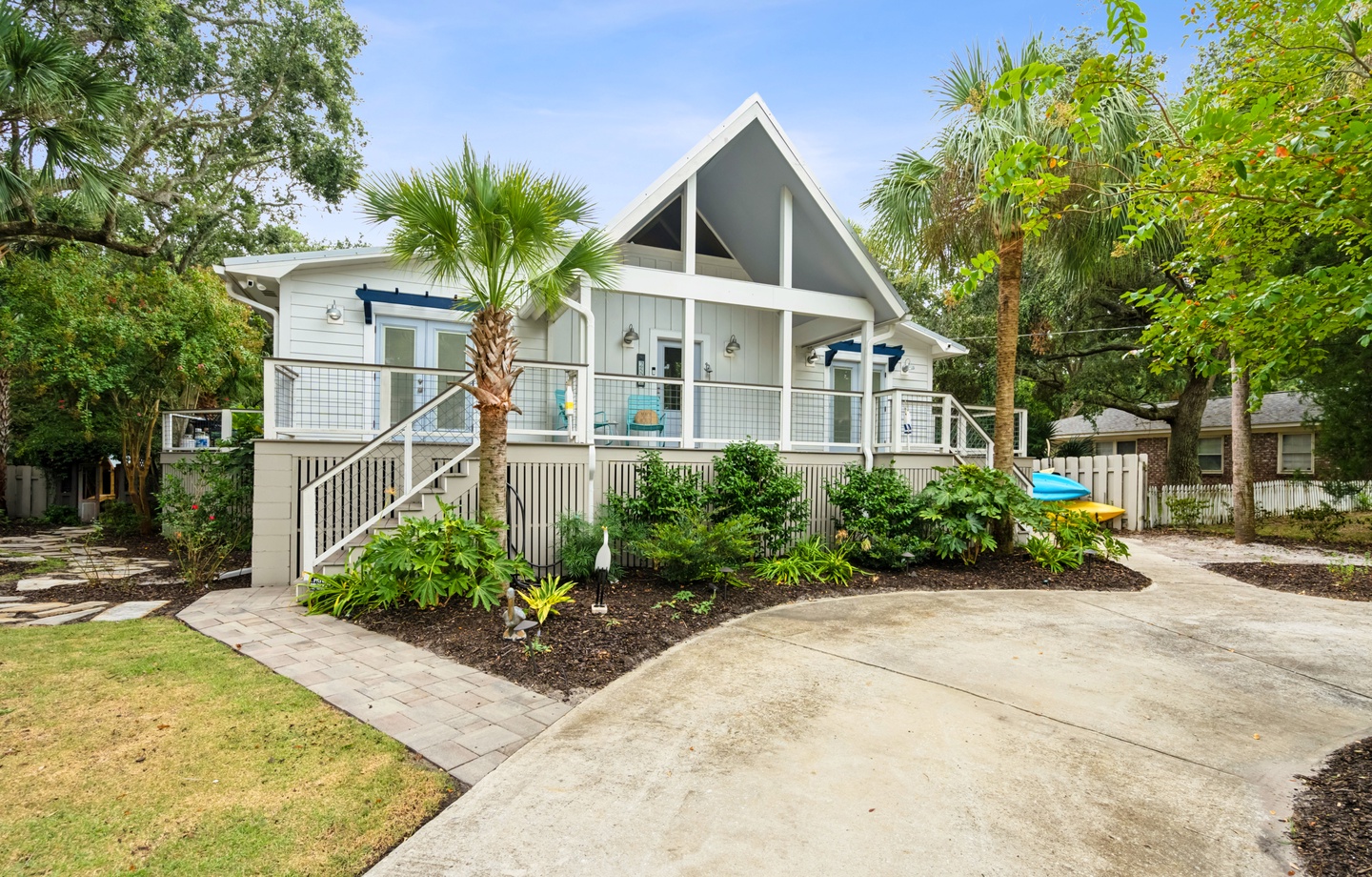 A charming coastal cottage with a peaked roof design, surrounded by lush tropical palms and native landscaping, welcoming guests to a peaceful retreat setting.