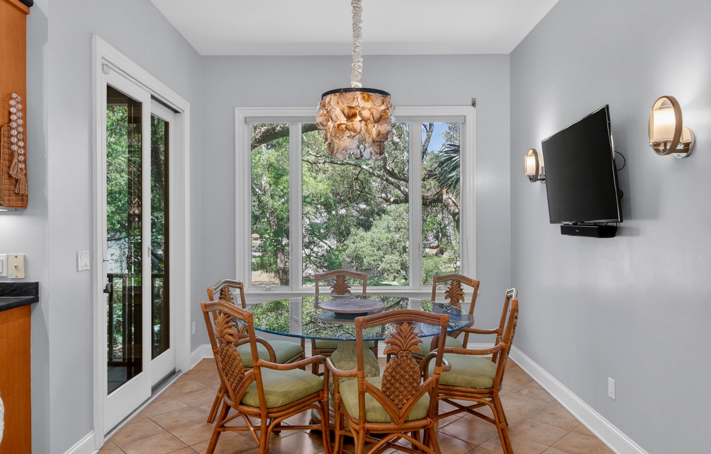 Gather around this inviting dining space with charming tropical-inspired seating and a stunning seashell chandelier, all bathed in natural light from large windows overlooking lush greenery.