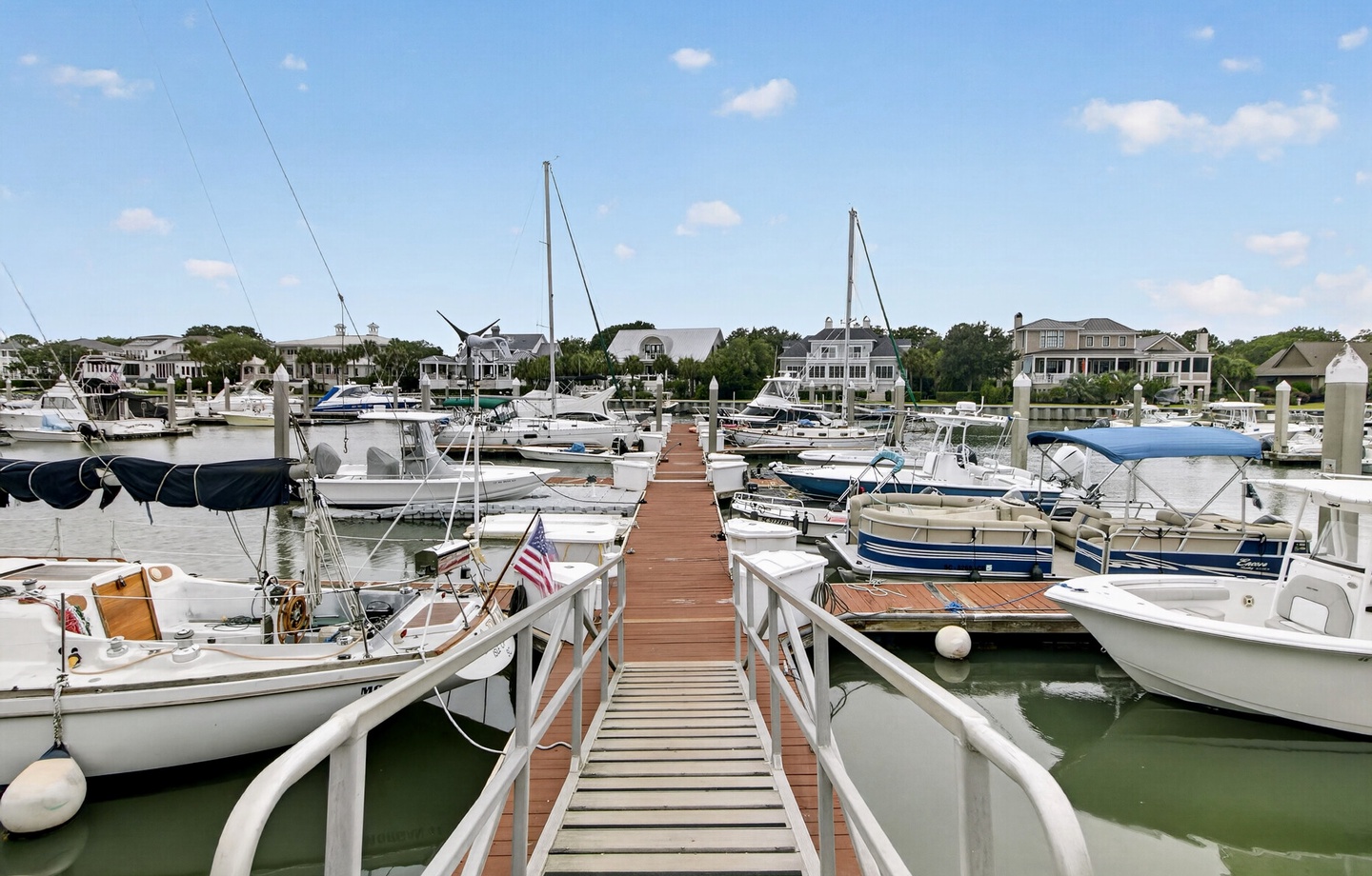 A picturesque marina with boats docked along a wooden pier, surrounded by waterfront homes and lush greenery under a clear blue sky.