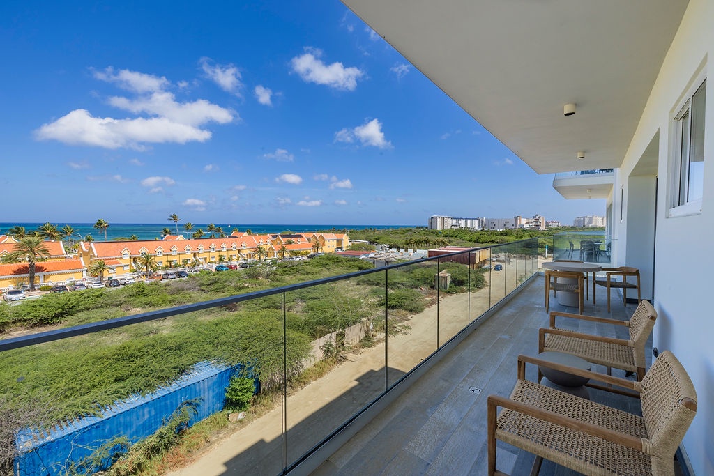 A modern balcony terrace offering sweeping sea views beyond the coastal neighborhood. Glass railings frame the turquoise horizon, while comfortable woven lounge chairs invite relaxation outdoors.