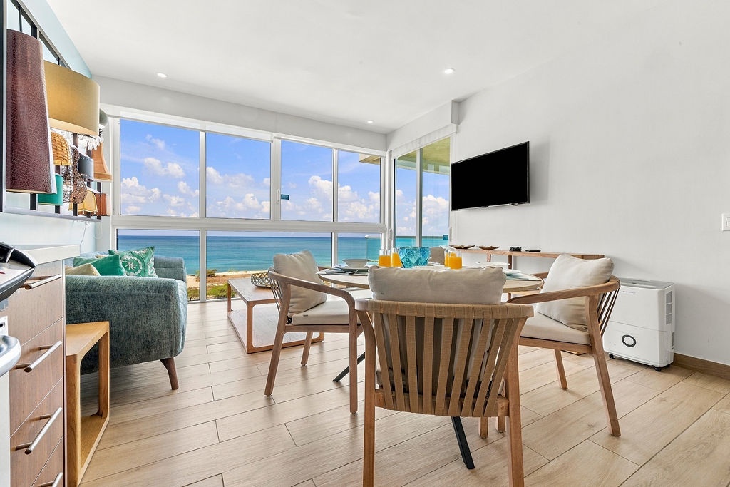 Gather around the warm wood dining table while natural light and stunning turquoise waters flood your open living space through floor-to-ceiling windows—your perfect backdrop for memorable meals and easy coastal living.