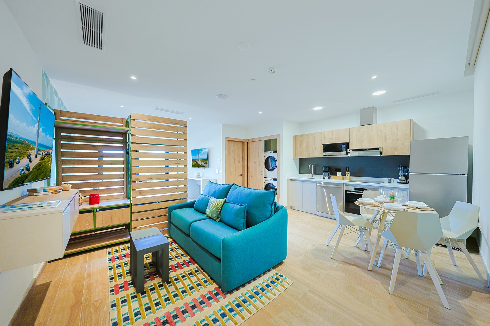 Picture yourself unwinding in this bright, modern space—vibrant turquoise sofa, fully-equipped kitchen, and cheerful dining nook ready for your seaside escape.