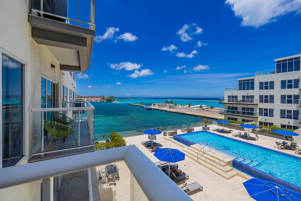 Modern oceanfront property features a pristine pool deck with stunning turquoise waters stretching to the horizon beyond adjacent buildings and manicured grounds.