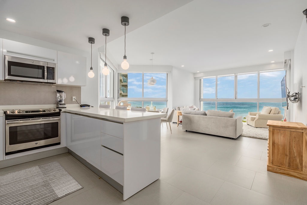 Your oceanfront retreat features a sleek white kitchen flowing into a bright living area with stunning sea views through floor-to-ceiling windows.