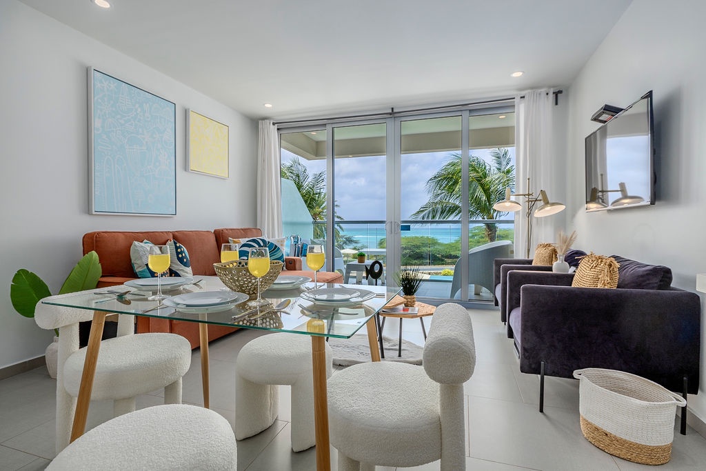 Relax in your bright modern living space where ocean breezes flow through floor-to-ceiling windows, revealing stunning sea views beyond swaying palms.