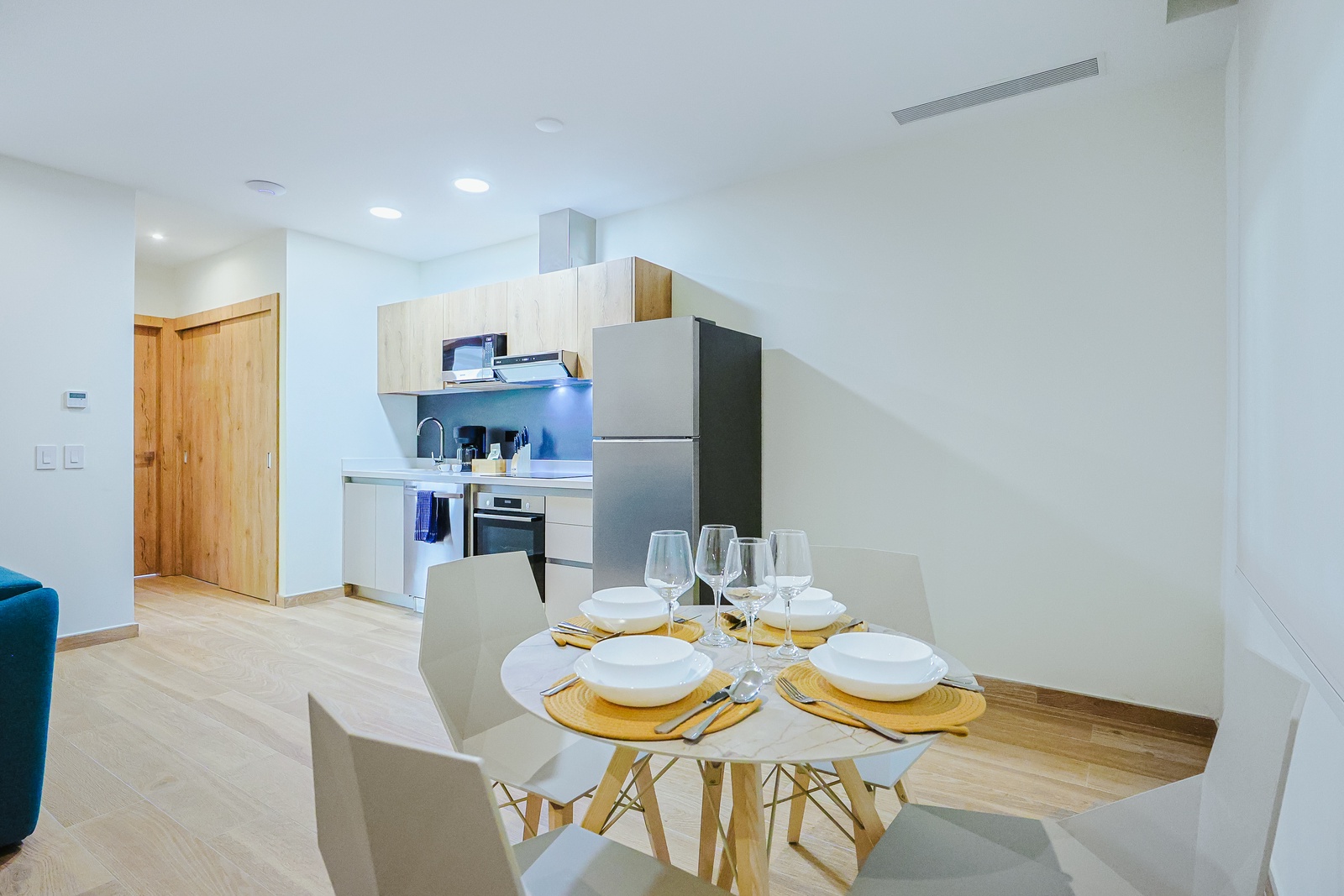 A bright, modern open-plan kitchen and dining space, elegantly set for four with golden placemats and fresh glassware. The sleek compact kitchen offers everything you need to prepare meals during your stay.