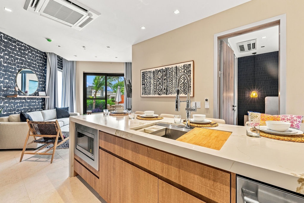 Step into your stylish open-plan kitchen and living area, where warm textures meet modern design. The island is ready for meal prep or casual dining, while tropical views through large glass doors invite the outdoors in.