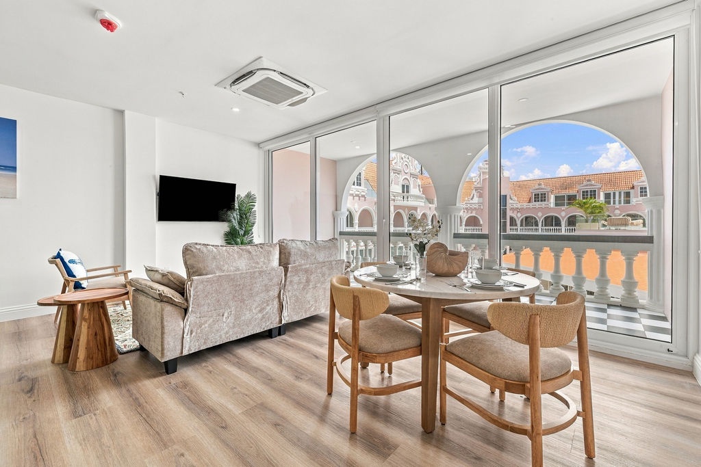 You'll love the bright, airy living space with floor-to-ceiling windows framing charming Mediterranean-style architecture. Relax on the cozy sofa or gather around the dining table for meals with that stunning backdrop.