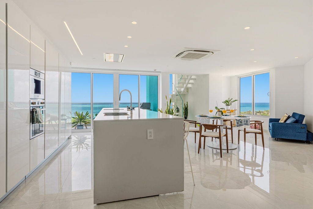 Step into this sunlit open-plan living space where ocean breezes meet modern style. Your kitchen flows seamlessly to the dining and lounge areas, all framed by breathtaking turquoise waters through floor-to-ceiling glass.
