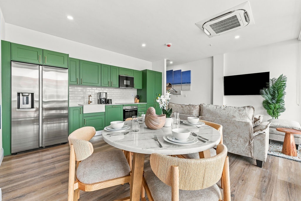Open-concept living and dining space with vibrant green cabinetry, stainless steel appliances, plush seating, and a marble dining table under soft recessed lighting.
