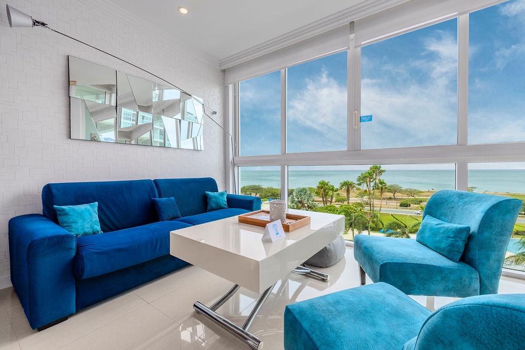 Relax into plush blue seating as panoramic windows frame stunning ocean views, palm trees, and coastal greens—your private retreat overlooking paradise.