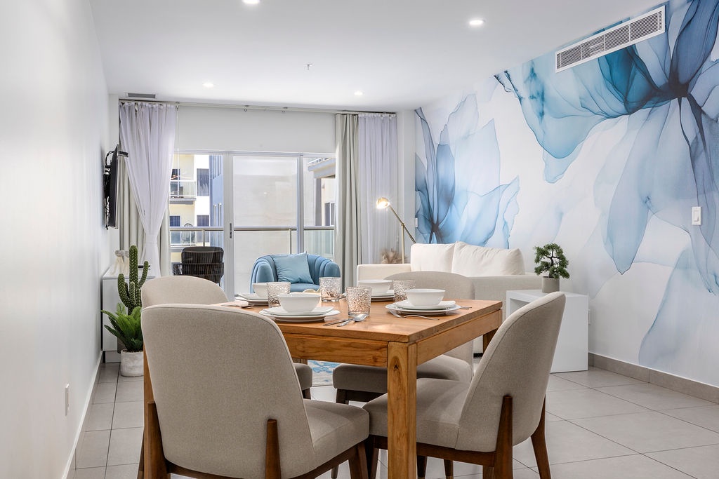 Gather around the warm wood table for memorable meals beneath calming blue floral art. Soft natural light spills through your private balcony, creating a serene space to unwind.