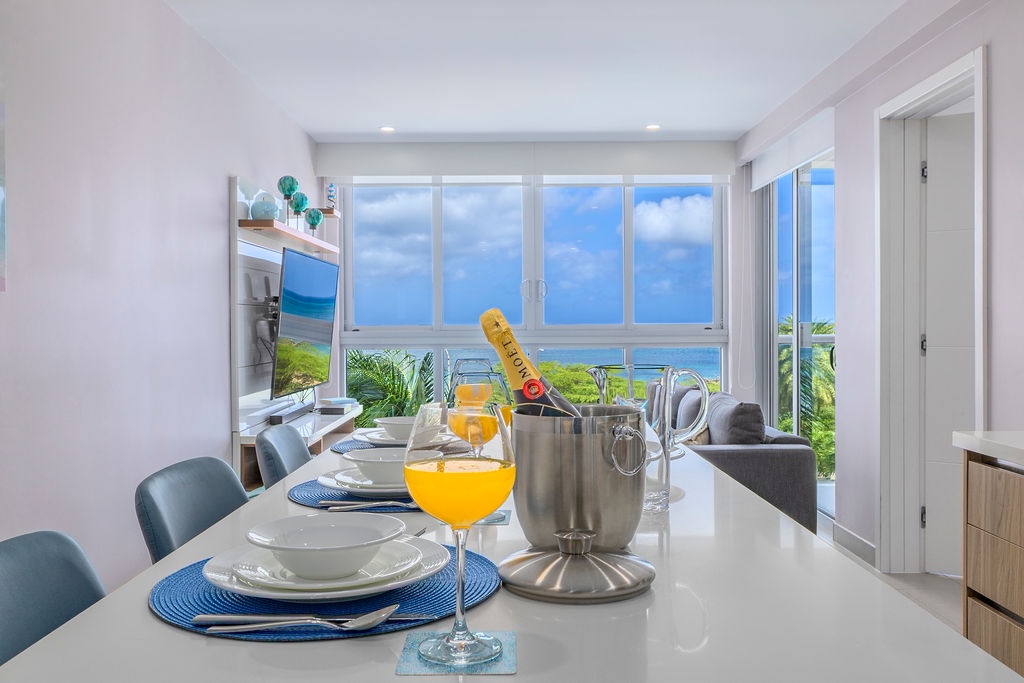 A bright, open dining space frames stunning ocean views through floor-to-ceiling windows, with champagne chilling and tropical breezes ready to welcome you home.