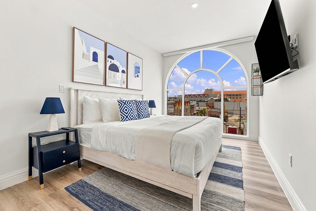Sink into soft linens and wake to the rooftops beyond your bright arched window. This serene bedroom blends crisp white tones with coastal blue accents, inviting you to relax and recharge in effortless comfort.