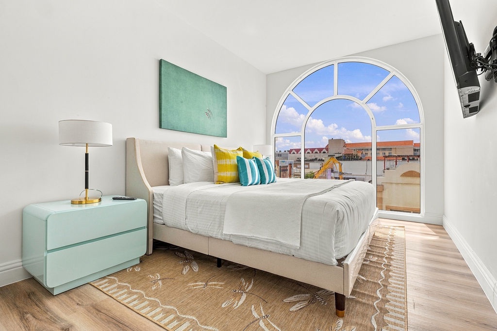 Your bright bedroom features a plush bed with fresh linens, colorful accent pillows, and a beautiful arched window framing city rooftops and blue sky—perfect for waking up to natural light.