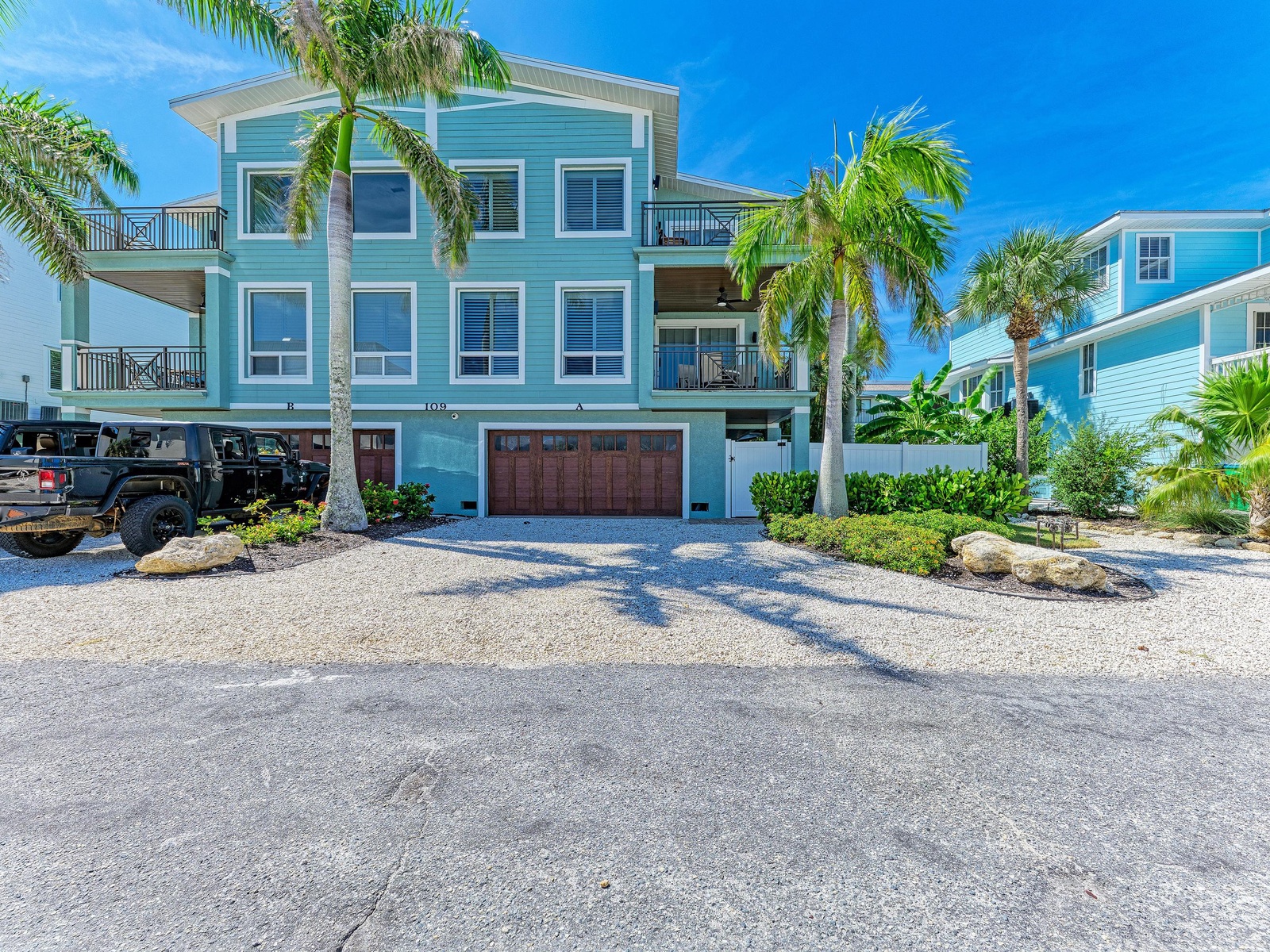 A coastal three-story property featuring modern Caribbean-style architecture, surrounded by lush palm trees and tropical landscaping, with convenient parking and beach access nearby.