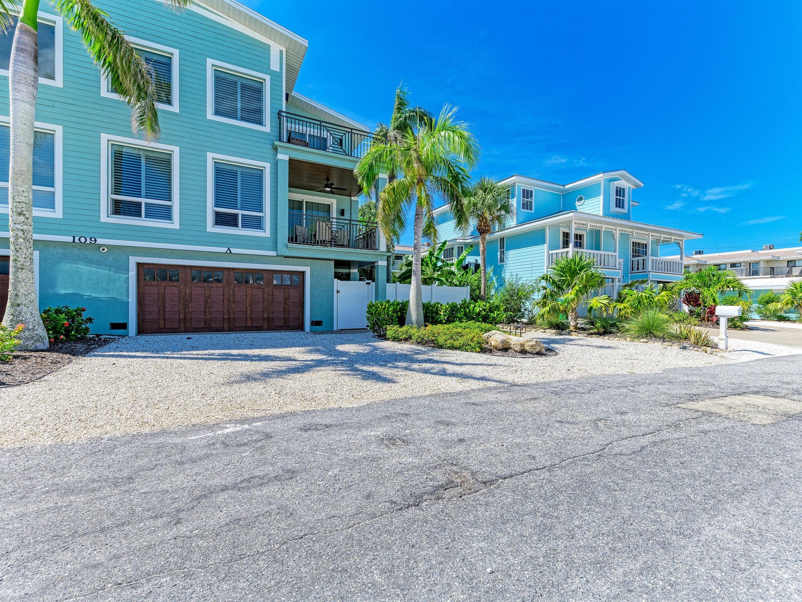 Coastal charm greets visitors as colorful beach homes line a quiet street, surrounded by swaying palms and lush tropical landscaping under clear blue skies.