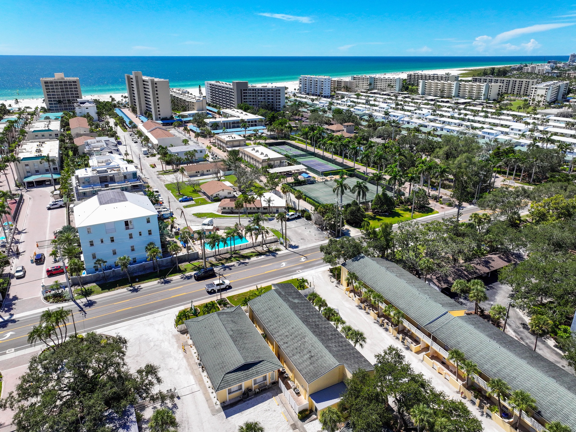 An aerial view showcasing a vibrant beachfront community with pristine white sands, turquoise waters, and the property nestled among palm-lined streets near tennis courts and resort amenities.