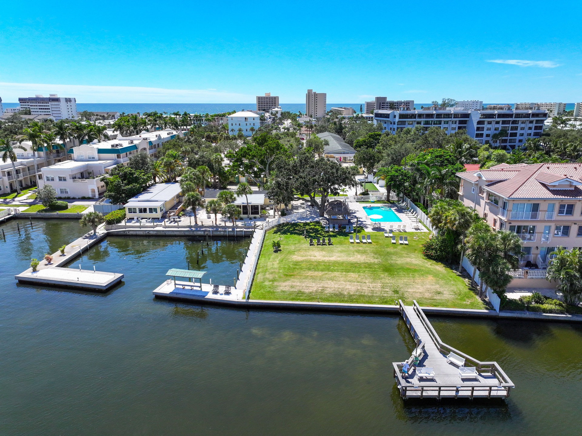 Aerial view of a waterfront vacation property featuring private docks, a sparkling pool, and lush grounds along a peaceful canal, with the beach just moments away.