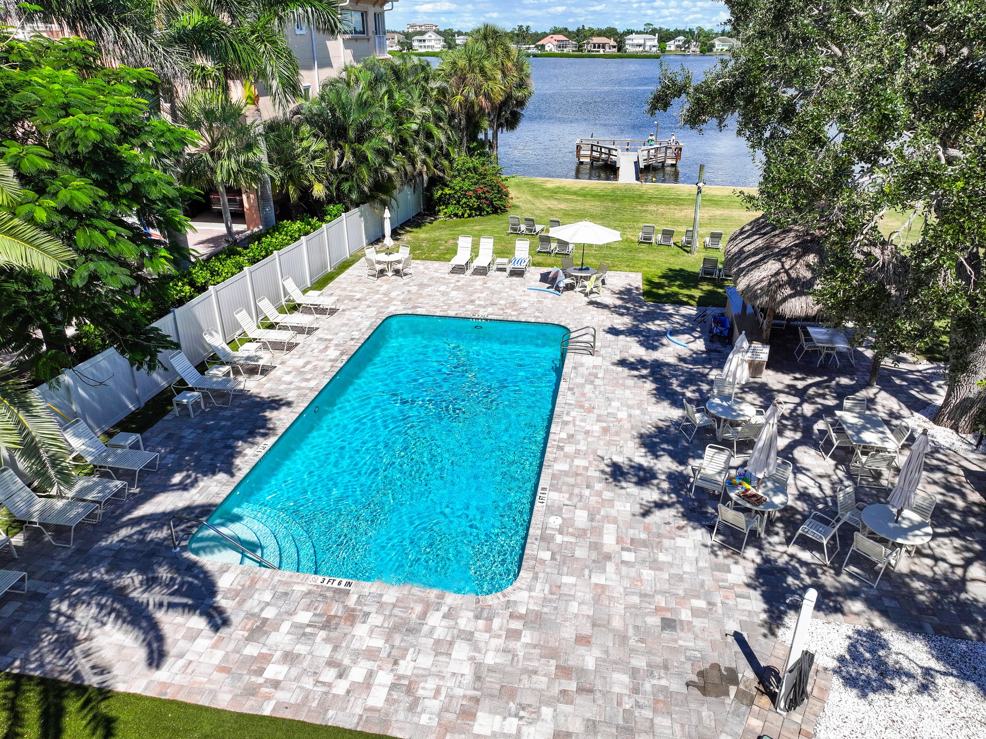 Aerial view of the property showcasing a sparkling outdoor pool surrounded by paver decking, lounge seating, and lush tropical landscaping. A private dock extends over calm lakefront waters, with neighboring homes visible across the shore.