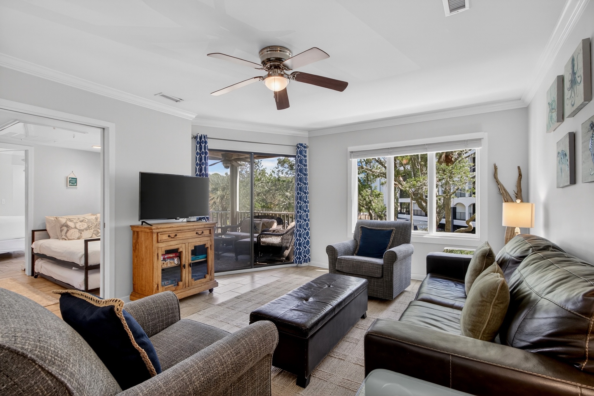 Unwind in this comfortable living room, where you can enjoy coastal breezes through the ceiling fan and abundant natural light from expansive windows framing lush greenery outside.