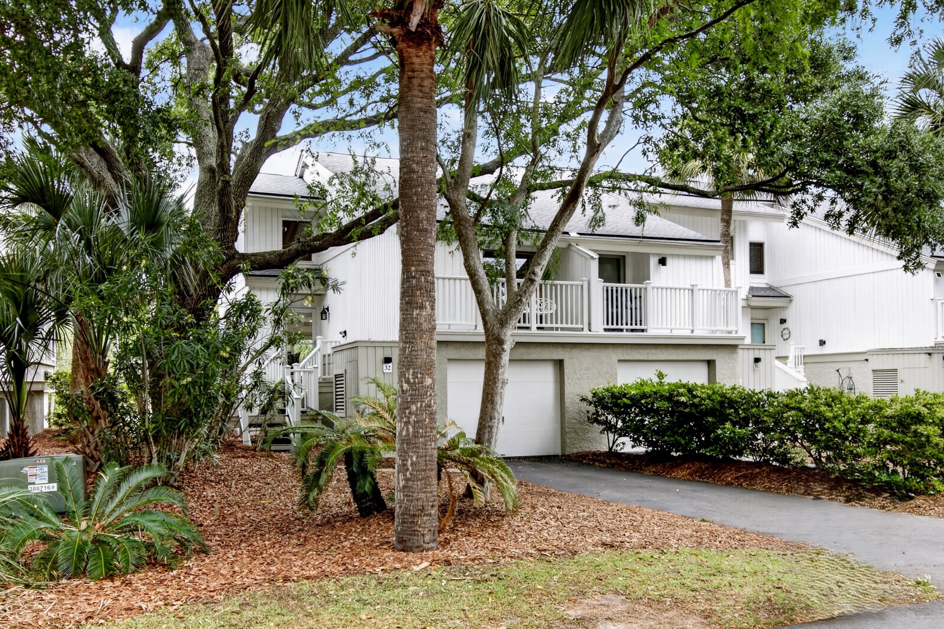 A coastal retreat nestled among lush tropical landscaping with palms and live oaks, featuring a private driveway leading to garage parking beneath covered balconies.