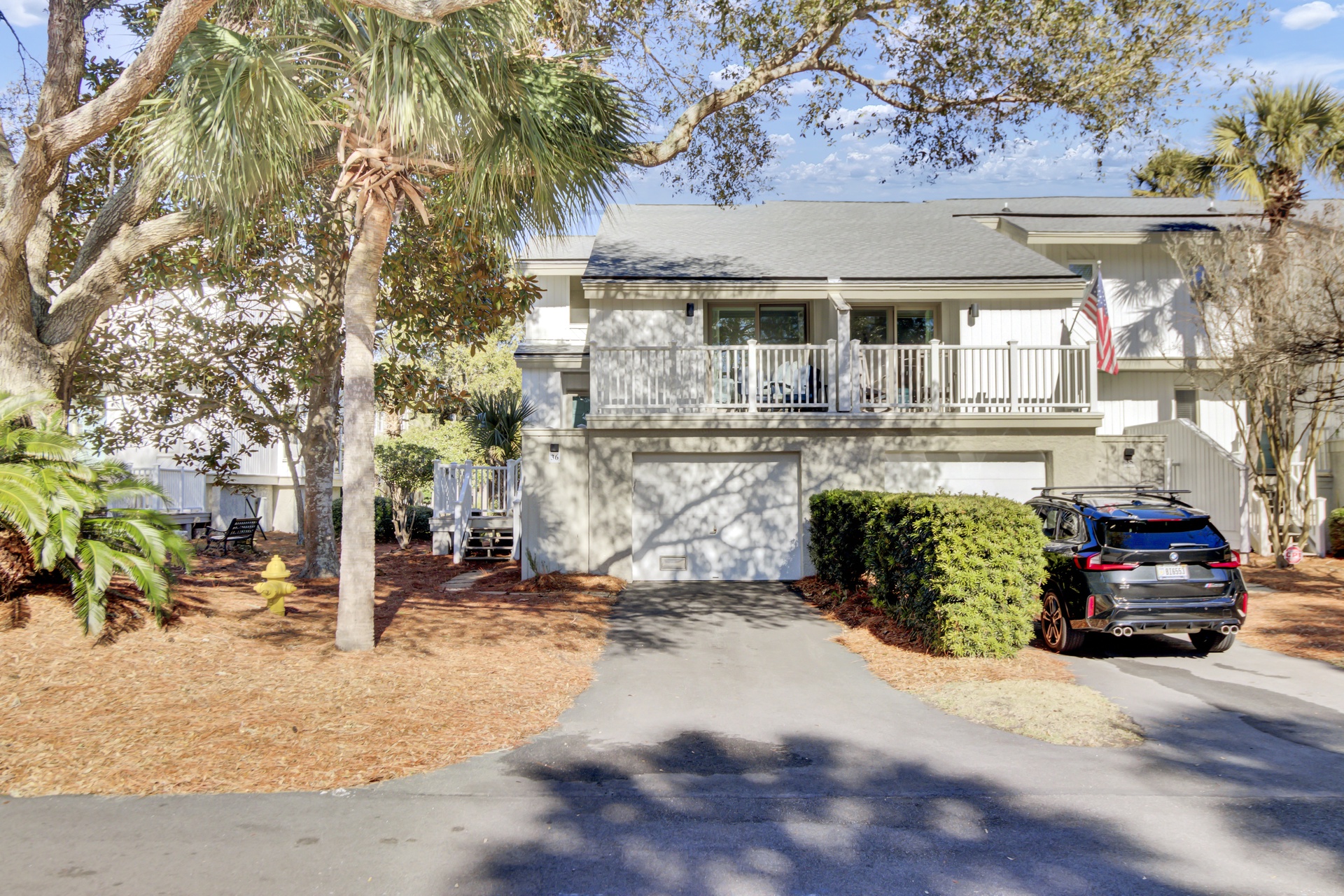 Charming two-story beach house with tropical palms and spacious balcony, featuring convenient parking and welcoming American flag display.