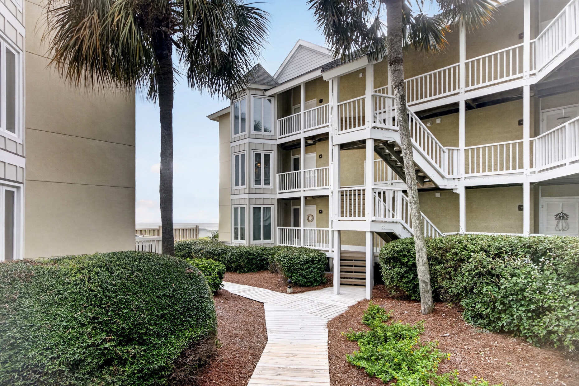 Coastal property buildings with white-railed balconies framing a sunny courtyard walkway, lined by palm trees and manicured hedges—your vacation getaway awaits.