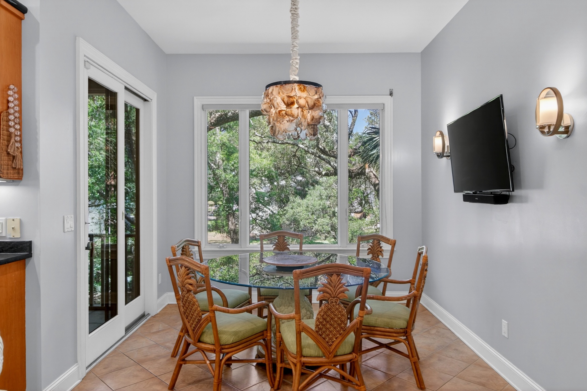 Gather around this inviting dining space with charming tropical-inspired seating and a stunning seashell chandelier, all bathed in natural light from large windows overlooking lush greenery.
