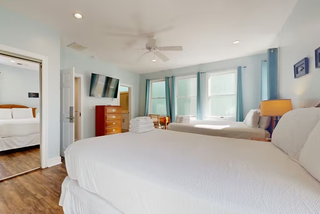 Unwind in your spacious bedroom featuring crisp linens, natural light through turquoise curtains, and modern comforts including a ceiling fan and flat-screen TV.
