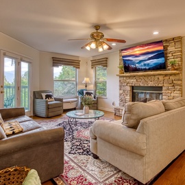 Unwind in your comfortable living room with a stone fireplace, cozy seating, and lovely natural light from multiple windows opening to peaceful greenery outside.