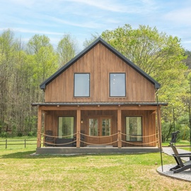 Modern wood-clad cabin nestled among trees with a welcoming covered porch and fire pit area perfect for evening gatherings.