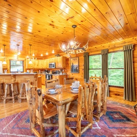 Gather around rustic log furniture in your open-concept cabin, where warm wood surrounds you and natural light streams through large windows overlooking peaceful greenery.