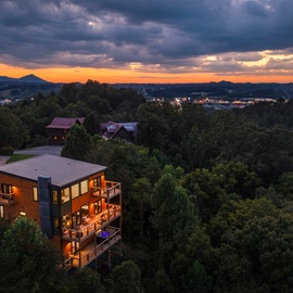This elevated mountain property sits surrounded by lush forest, with the town of Pigeon Forge glowing softly in the valley below and dramatic sunset colors painting the sky.