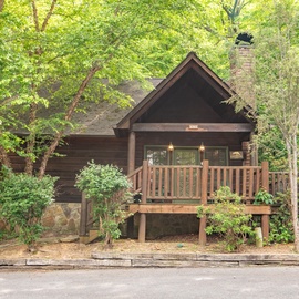 A charming log cabin nestled among lush trees, offering a cozy woodland retreat. The covered front porch and natural stone accents welcome guests to this peaceful mountain escape.