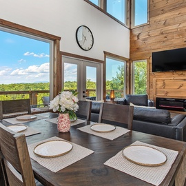 Gather around your private dining table where expansive windows frame stunning forest vistas. Sunlight streams through floor-to-ceiling glass, highlighting the warm wood details and seamless connection to your outdoor deck.