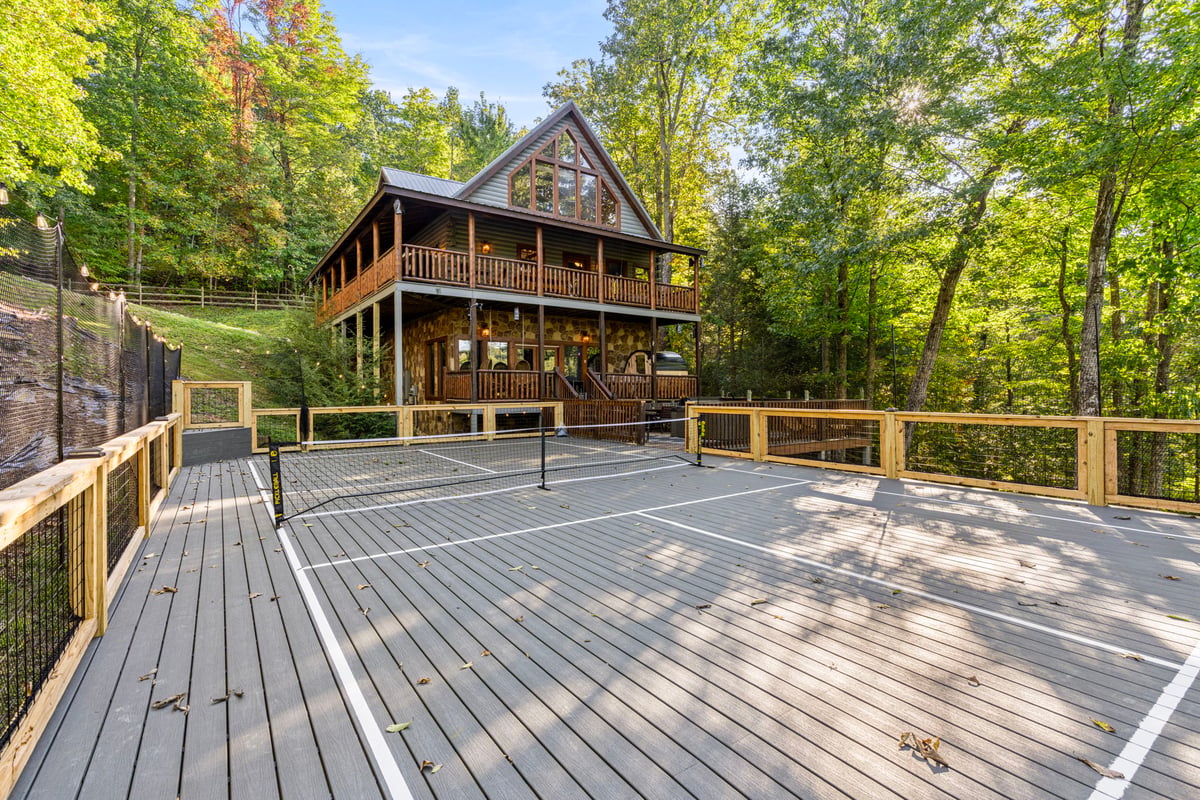 A charming A-frame cabin nestled among tall trees, featuring wrap-around balconies and a pickleball court perfect for outdoor games. The property offers a private woodland setting with natural beauty all around.