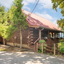 Charming log cabin with a rustic metal roof nestled among tall trees, offering a secluded mountain retreat with a welcoming front porch and easy parking access.